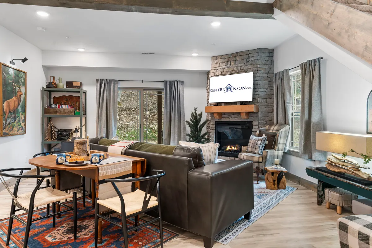 The image showcases a spacious, inviting interior with a glowing electric fireplace as the focal point, surrounded by cozy seating. Large windows flood the room with natural light, and a welcoming dining table is perfectly situated for family meals or game nights.