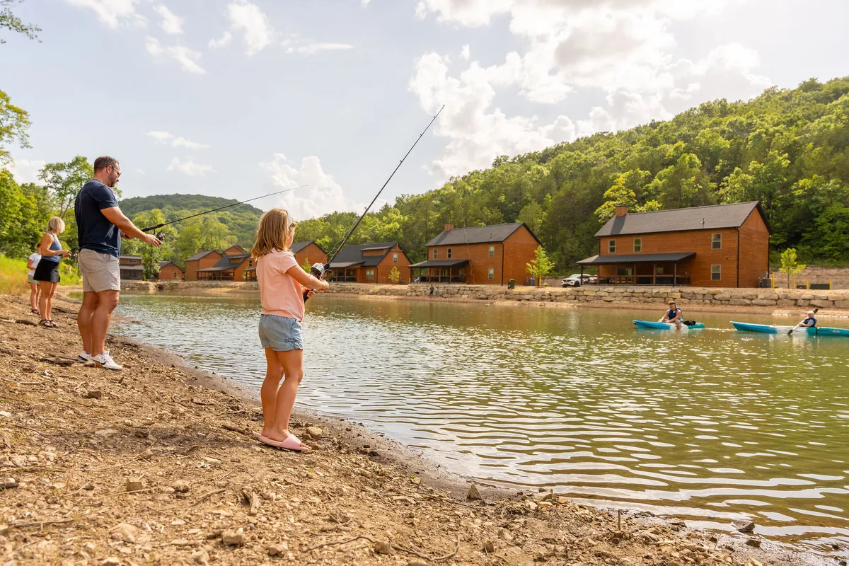 A family is fishing by a serene lake, with kayakers enjoying the calm water and rustic cabins nestled in the background. The scene captures an active yet tranquil day outdoors, with plenty of greenery and a playful, inviting mood.