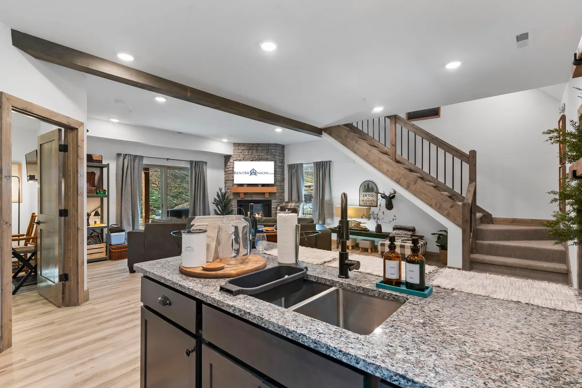 A bright interior showcases a kitchen island with bar seating, a staircase leading upstairs, a fireplace, and expansive windows that flood the area with sunlight. The layout encourages fluid movement between kitchen and lounge for effortless entertaining.