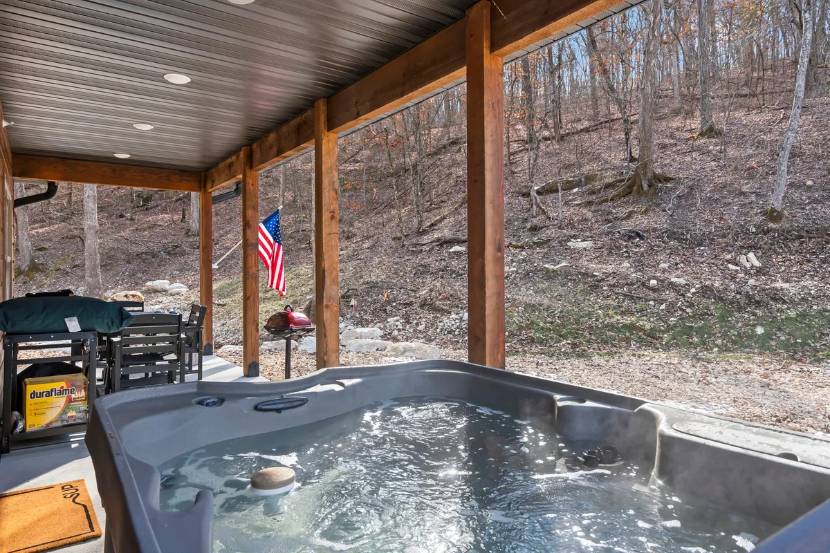 The outdoor area features a bubbling hot tub, inviting seating, and an American flag on a covered porch. The scene evokes relaxation, with elements like the hot tub and patio reflecting private resort-style comforts.