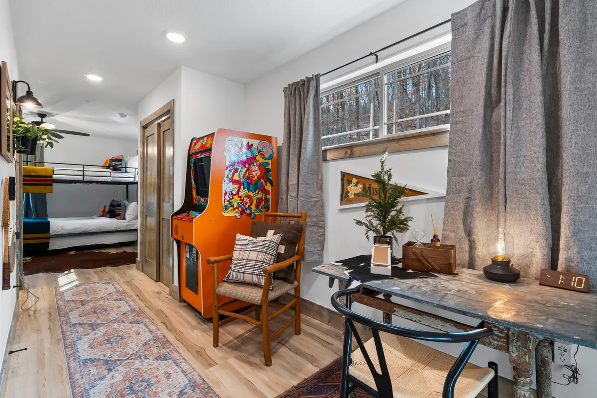 A playful bunk room setting with a bunk bed, an arcade machine, a wooden desk, a decorative plant, and cozy textiles on the floor. The space feels fun, creative, and perfectly suited for children or teens.