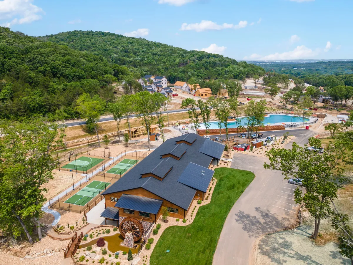 A lively view of the main resort building, with a sparkling swimming pool in the foreground, multiple sports courts, and beautifully landscaped gardens. The area is bathed in sunlight, hinting at energetic social gatherings and recreational play.