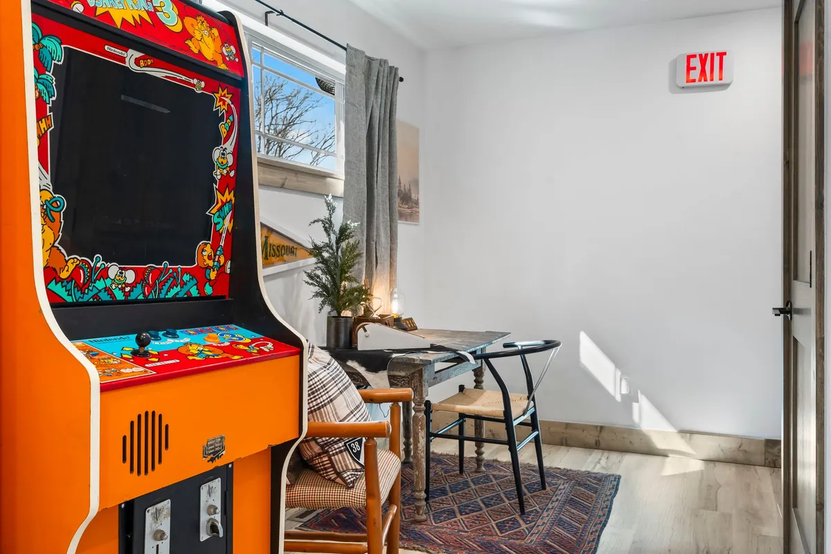 This image features an inviting interior space with a classic arcade machine, a wooden table, a decorative plant, and a sunlit window with curtains. The scene radiates a fun and laid-back vibe, suggesting a spot for both competitive gaming and relaxed socializing.