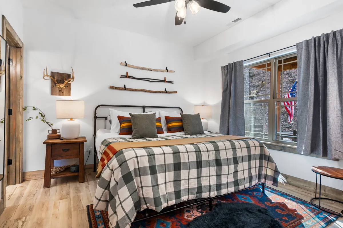 A cozy main-level bedroom featuring a bed with plaid bedding, rustic decor elements, and a window with curtains. The room is bathed in natural light, offering a welcoming and peaceful atmosphere.