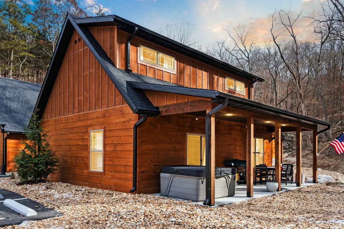 This exterior shot features the cabin's wooden facade, a private hot tub, a cozy seating area on the patio, and the American flag. The surrounding greenery creates a comfortable and inviting atmosphere, perfect for relaxing after work or connecting with nature.