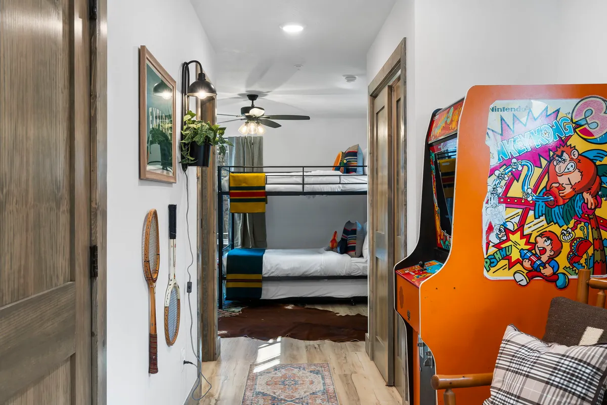 A cozy bunk room featuring a bunk bed, an arcade game, decorative wall art, and wooden flooring. The space is designed for fun and relaxation, with elements that appeal to both children and families gathering for games or bedtime stories.