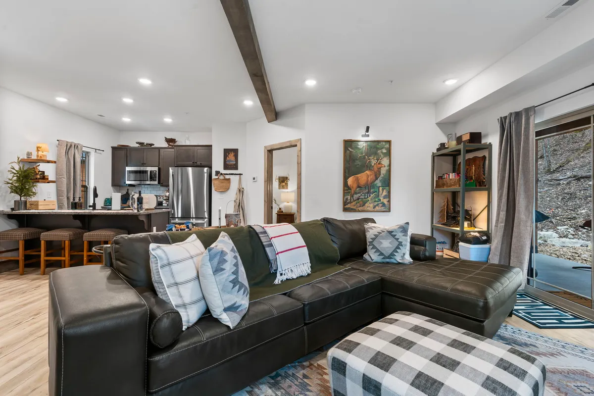 A cozy living room features a large leather sectional sofa adorned with plaid throw blankets, a modern electric fireplace as the focal point, framed deer artwork above the mantel, and bar stools at the kitchen counter that hint at the open-concept layout. The space is bathed in natural light, creating a welcoming atmosphere ideal for gatherings.