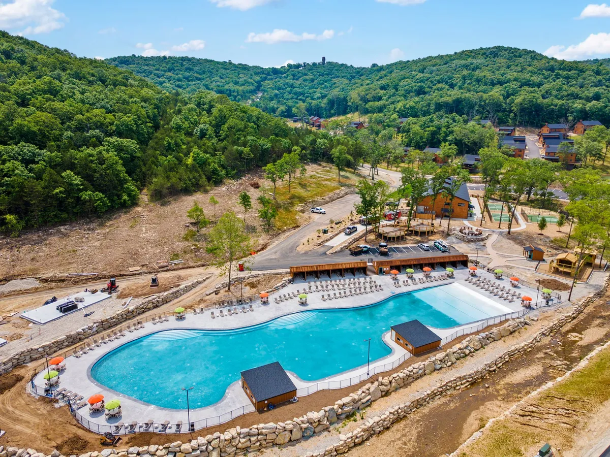 A large outdoor swimming pool bordered by sun loungers and greenery, with playground equipment visible in the background. The inviting pool area emphasizes opportunities for swimming, sunbathing, and outdoor family fun.